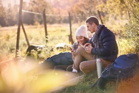 Junges Wanderpärchen, das sich im sonnigen Gras ausruht und dabei ein Mobiltelefon und ein digitales Tablet benutzt, lizenzfreies Stockfoto