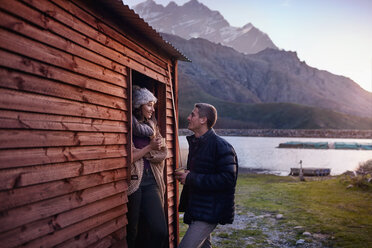 Junges Paar trinkt Kaffee an der Tür einer Hütte am See - CAIF05083