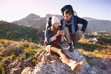 Young couple with backpacks hiking, resting on rock using smart phone - CAIF05085