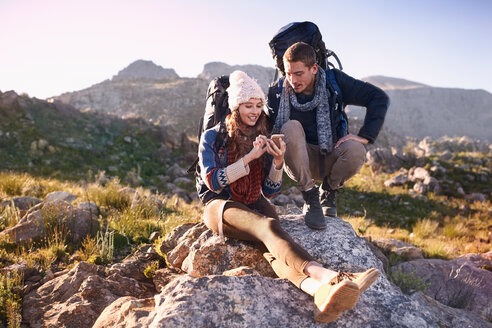 Junges Paar mit Rucksäcken beim Wandern, ruht sich auf einem Felsen aus und benutzt ein Smartphone - CAIF05085
