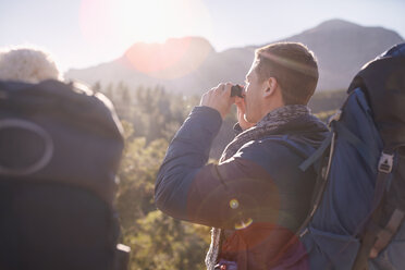 Young man with backpack hiking using binoculars in sunny field - CAIF05090