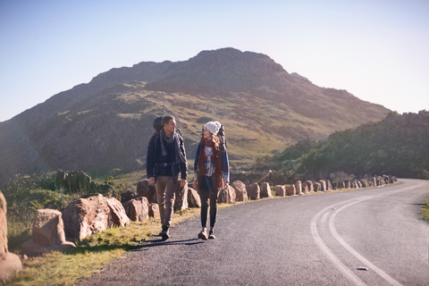 Junges Paar mit Rucksäcken beim Wandern entlang einer abgelegenen, sonnigen Straße, lizenzfreies Stockfoto