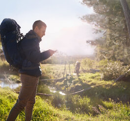 Young man with backpack hiking, checking smart phone in sunny field - CAIF05103