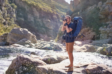 Junger Mann mit Rucksack wandernd, fotografierend mit Kamera auf sonnigen Felsen - CAIF05107