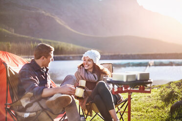 Lächelndes junges Paar trinkt Kaffee auf einem sonnigen Campingplatz am See - CAIF05118