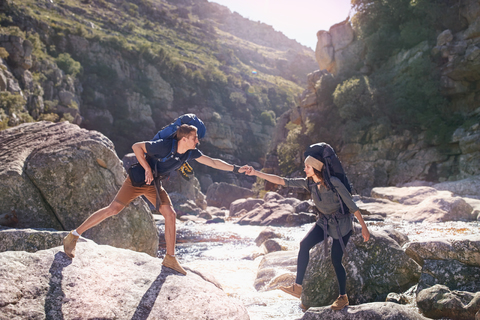 Junger Mann hilft Freundin beim Wandern, überquert sonnige Felsen im Bach, lizenzfreies Stockfoto