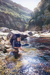 Young woman with backpack hiking, washing hands at sunny stream - CAIF05133