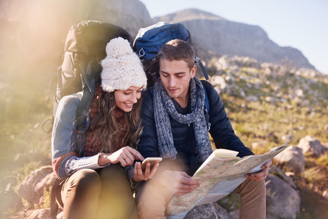 Junges Paar mit Rucksäcken beim Wandern, rastend und auf Karte und Smartphone schauend, lizenzfreies Stockfoto