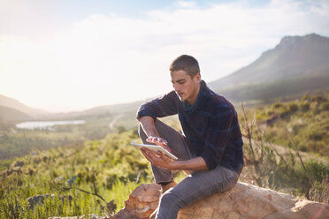 Junger Mann mit digitalem Tablet auf einem Felsen in einem sonnigen, abgelegenen Tal - CAIF05141