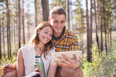 Junges Paar zeltet, trinkt Kaffee und benutzt ein digitales Tablet in einem sonnigen Wald - CAIF05152