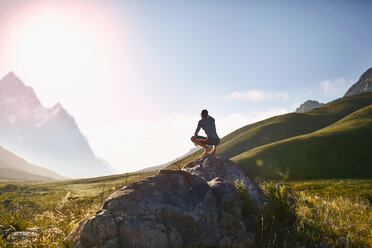 Junger Mann kauert auf einem Felsen und blickt auf einen sonnigen, stimmungsvollen Bergblick - CAIF05155