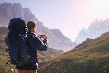 Junger Mann mit Rucksack und Fotohandy in einem sonnigen Tal unterhalb der Berge - CAIF05156