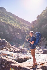 Junger Mann mit Rucksack beim Wandern und Fotografieren von sonnigen, zerklüfteten Felsen mit Kamera - CAIF05161