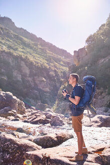 Young man with backpack hiking and photographing sunny, craggy cliffs with camera - CAIF05161