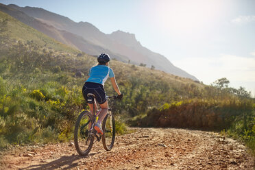 Young woman mountain biking on sunny, remote dirt road - CAIF05163