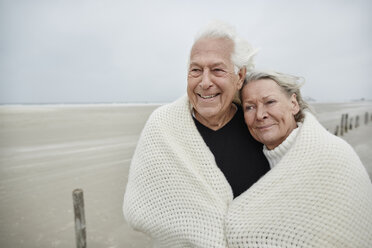 Lächelndes, liebevolles, älteres Paar, eingewickelt in eine Decke am Strand - CAIF05170