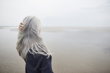 Ältere Frau mit Hand im langen grauen Haar am Winterstrand - CAIF05171