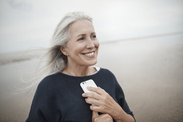 Smiling senior woman holding cell phone on winter beach - CAIF05173