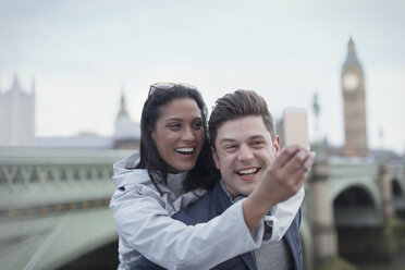 Playful, affectionate couple tourists taking selfie with camera phone in front of Westminster Bridge, London, UK - CAIF05194