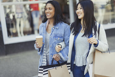 Lächelnde Freundinnen, die mit Kaffee und Einkaufstüten Arm in Arm an einer Ladenfront entlang gehen - CAIF05255