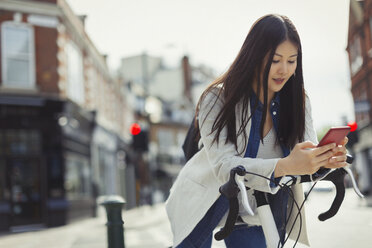 Young woman commuting on bicycle, texting with cell phone on sunny urban street - CAIF05259