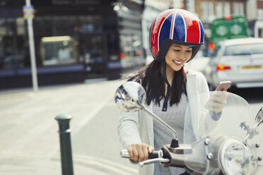 Lächelnde junge Frau, die auf einem Motorroller mit Handy SMS schreibt und einen Helm auf einer städtischen Straße trägt - CAIF05264