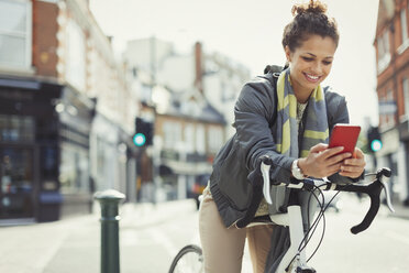Lächelnde junge Frau, die mit dem Fahrrad unterwegs ist und auf einer sonnigen städtischen Straße mit dem Handy telefoniert - CAIF05265
