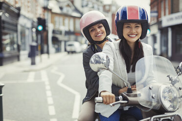 Smiling young women friends wearing helmets, riding motor scooter on urban street - CAIF05276