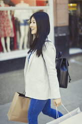 Smiling young woman walking along storefront with shopping bags - CAIF05288