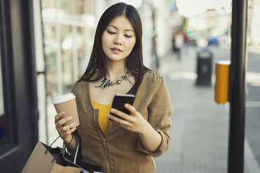 Junge Frau mit Kaffee, die auf dem Bürgersteig mit ihrem Handy SMS schreibt - CAIF05291