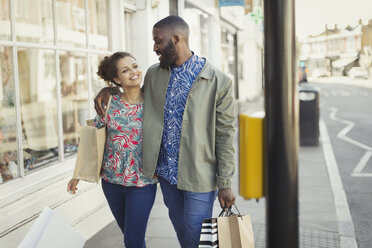 Verliebtes junges Paar mit Einkaufstüten, das an einer städtischen Ladenfront entlanggeht - CAIF05295