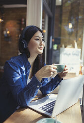 Pensive young woman listening to music with headphones and drinking coffee at laptop in cafe window - CAIF05298