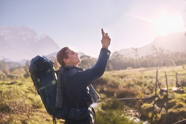 Junger Mann mit Rucksack beim Wandern und mit Fotohandy in einem sonnigen, abgelegenen Feld - CAIF05304