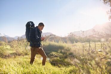 Young man with backpack hiking, checking compass in sunny, remote field - CAIF05305