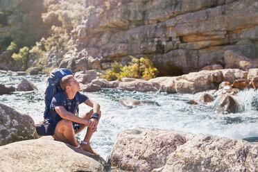 Junger Mann mit Rucksack beim Wandern, rastend an einem sonnigen, felsigen Bach - CAIF05306