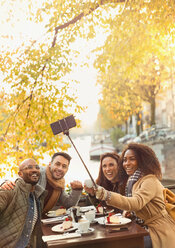 Lächelnde Freunde, die ein Selfie mit einem Selfie-Stick im Herbst in einem Straßencafé machen - CAIF05376