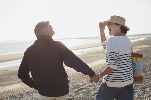 Lachendes reifes Paar, das sich an den Händen hält und am sonnigen Strand spazieren geht, lizenzfreies Stockfoto
