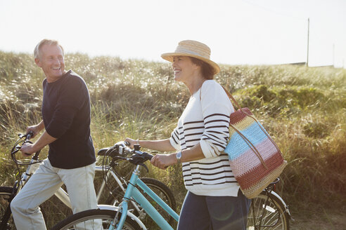 Älteres Paar, das auf einem sonnigen Grasweg am Strand Rad fährt - CAIF05393