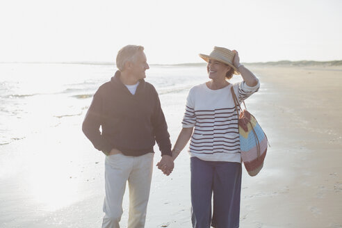 Lächelndes reifes Paar, das sich an den Händen hält und am sonnigen Strand spazieren geht - CAIF05396