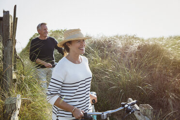 Älteres Paar geht mit Fahrrädern am sonnigen Strand entlang - CAIF05399