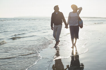 Affectionate mature couple holding hands and walking on sunset ocean beach surf - CAIF05411