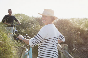 Portrait smiling mature woman walking bicycle on sunny beach grass path - CAIF05416