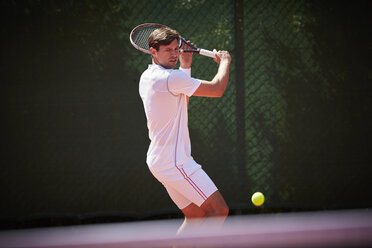 Junger männlicher Tennisspieler, der auf einem sonnigen Tennisplatz den Tennisball schwingt - CAIF05480