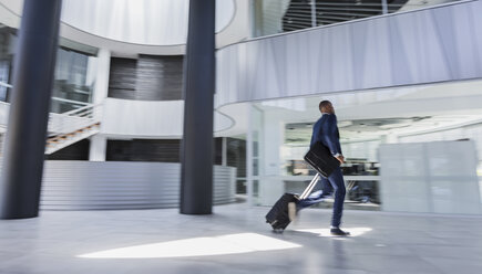 Businessman rushing, pulling suitcase in modern office lobby - CAIF05529