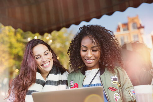 Smiling young women friends using laptop at sidewalk cafe - CAIF05643