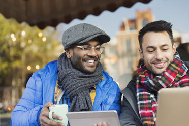 Lächelnde junge Männer, die in einem Straßencafé Kaffee trinken und einen Laptop und ein digitales Tablet benutzen - CAIF05646