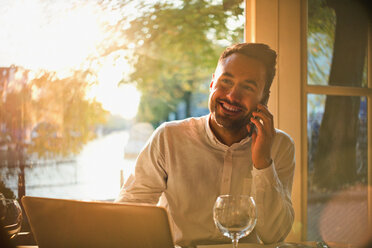 Lächelnder junger Mann, der mit seinem Handy telefoniert und einen Laptop in einem sonnigen Café benutzt - CAIF05651