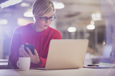 Seriöse Geschäftsfrau, die bis spät in die Nacht arbeitet und ein Mobiltelefon am Laptop im Büro benutzt - CAIF05695