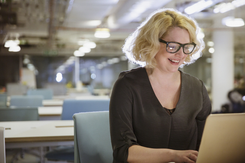 Lächelnde Geschäftsfrau, die spät am Laptop in einem dunklen Büro arbeitet, lizenzfreies Stockfoto