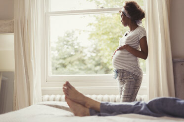 Nachdenkliche schwangere Frau schaut aus dem Schlafzimmerfenster - CAIF05711